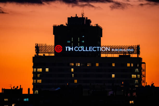 La imagen muestra uno de los hoteles de la cadena NH que hay en Madrid. La imagen está tomada al atardecer viéndose un cielo anaranjado.