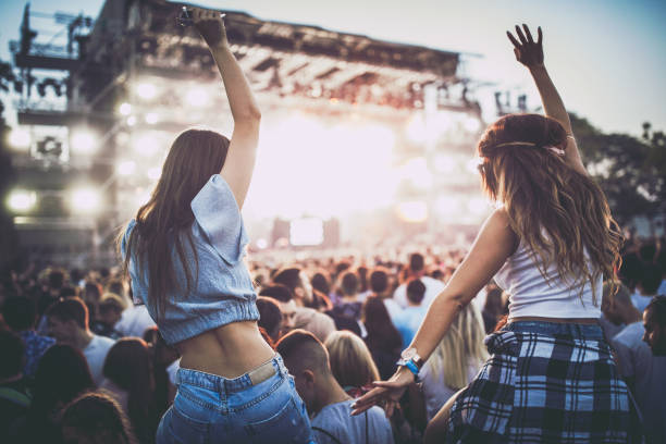 En la imagen se puede ver como protagonista de la imagen dos chicas subidas a hombros pasándolo bien en un concierto o festival, se puede ver que donde están hay mucha gente y el escenario lleno de luces.