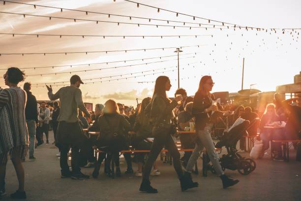 En la imagen se puede ver un recinto de lo que parece ser un festival, al aire libre con puestos de comida y bebida y hay gente sentada en las mesas que hay para descansar.
