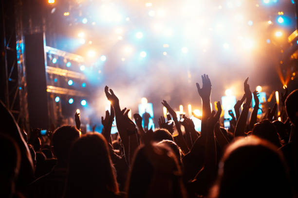 En la fotografía se ve una multitud en frente de un escenario, se puede ver que es de noche y están ambientados con unas luces de muchos colores. 